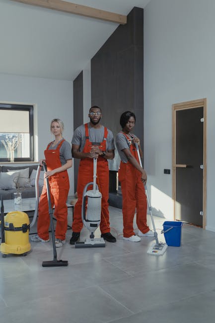 Three professional cleaners from Cleaners St. John's Wood, dressed in red overalls, are performing surface cleaning in a modern living room with natural light. The woman on the left holds a vacuum cleaner, while the woman on the right uses a mop and bucket, and the man in the center holds a small cleaning tool, all demonstrating thorough domestic cleaning. The room features a tiled floor, a grey wall with wooden accents, a large window with sunlight streaming in, and minimal furniture, emphasizing cleanliness and hygiene. Visible cleaning tools and equipment suggest a deep cleaning or sanitisation process aimed at maintaining a pristine environment, aligning with the services offered on the Price guide for house cleaning in St Johns Wood page by [COMPANY_NAME].