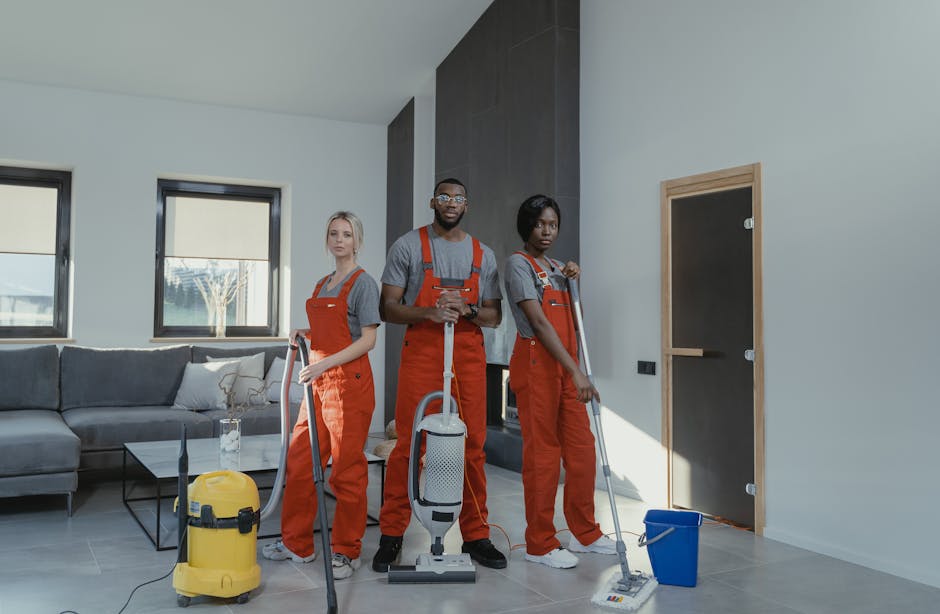 Three professional cleaners from Cleaners St. John's Wood are standing in a modern living room during a domestic cleaning session. They are dressed in gray T-shirts with red overalls and are equipped with cleaning tools including a vacuum cleaner, a mop, and a bucket. The room features a light-colored tiled floor which is being surface cleaned, with a dark gray sofa, cushions, and a glass coffee table visible in the background. The living room has large windows with blinds allowing natural light to illuminate the space, showcasing the neat and well-maintained surfaces. The cleaners are positioned near the door, ready to perform deeper cleaning and sanitisation of the area, reflecting the company’s expertise in detailed domestic cleaning services offered in the guide for house cleaning in St Johns Wood.