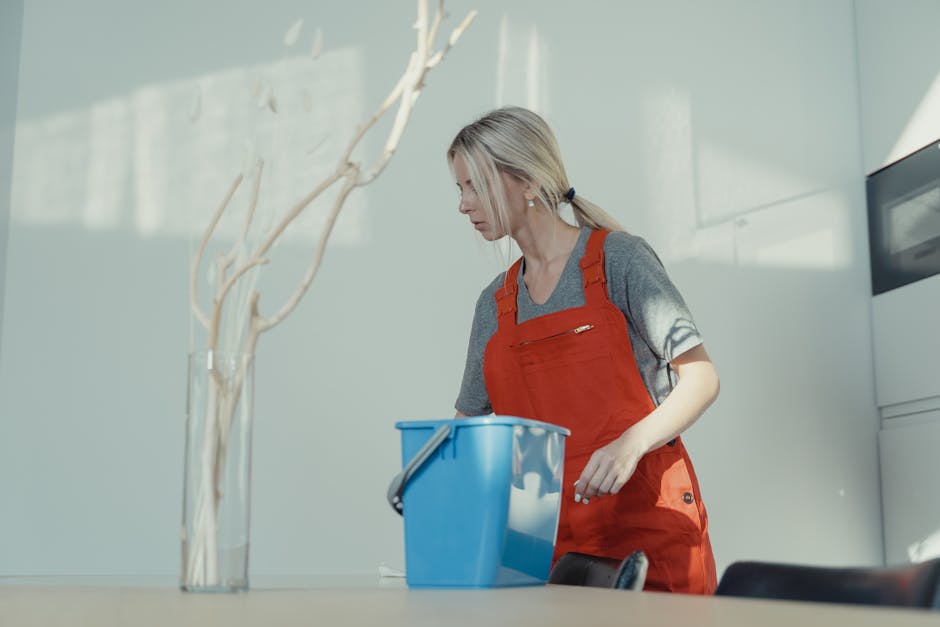Three professional cleaners from Cleaners St. John's Wood, dressed in red overalls, are performing surface cleaning in a modern living room with natural light. The woman on the left holds a vacuum cleaner, while the woman on the right uses a mop and bucket, and the man in the center holds a small cleaning tool, all demonstrating thorough domestic cleaning. The room features a tiled floor, a grey wall with wooden accents, a large window with sunlight streaming in, and minimal furniture, emphasizing cleanliness and hygiene. Visible cleaning tools and equipment suggest a deep cleaning or sanitisation process aimed at maintaining a pristine environment, aligning with the services offered on the Price guide for house cleaning in St Johns Wood page by [COMPANY_NAME].