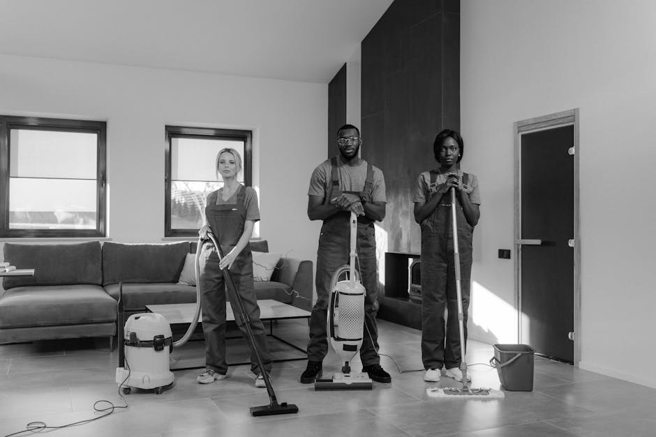Three professional cleaners from Cleaners St. John's Wood are engaged in surface cleaning and deep cleaning activities inside a modern residential living room. The room features a wooden floor that appears freshly vacuumed, with a polished, shiny surface free of dust and debris. The area includes a large, contemporary sofa with fabric upholstery, positioned beneath two sizeable windows allowing natural light to illuminate the space. The cleaners are dressed in work uniforms and are equipped with cleaning tools: the person on the left is operating a cylinder vacuum cleaner, the one in the middle is holding a portable unit, and the individual on the right is using a mop and bucket for floor sanitisation. The room’s décor includes a dark wood feature wall and a glass-fronted cabinet, contributing to a clean and well-maintained atmosphere. This image demonstrates professional domestic cleaning services offered by [COMPANY_NAME], ensuring hygienic and spotless living environments in line with end of tenancy cleaning standards.
